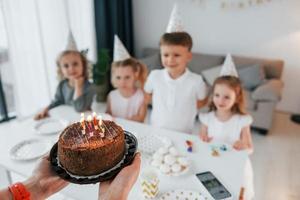 Frau, die Kuchen mit Kerzen hält. Geburtstag feiern. Gruppe von Kindern ist tagsüber zusammen zu Hause foto