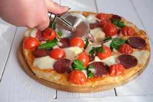 menschliche hand mit pizzaschneider schnitt pizza in der küche. foto