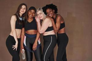 das Zusammensein genießen. Gruppe multiethnischer Frauen, die im Studio vor braunem Hintergrund stehen foto