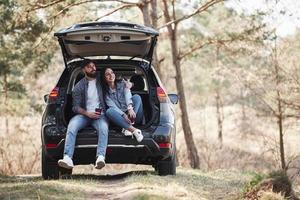 warme Atmosphäre. Sitzen auf dem hinteren Teil des Autos. die Natur genießen. Paar ist mit ihrem brandneuen schwarzen Auto im Wald angekommen foto