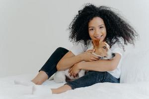 Foto einer hübschen jungen Tierhalterin posiert in einem sauberen weißen Schlafzimmer, umarmt den Hund, spielt mit dem besten Freund, trägt Freizeitkleidung, hat einen fröhlichen Ausdruck. menschen, tiere, liebe, freundschaftskonzept