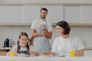 Glückliche Mutter spricht mit Tochter beim Frühstück. vater steht hinter, zubereitetes leckeres gericht für die familie. freundliche Familienmitglieder treffen sich am Wochenende in der Küche, genießen nette Gespräche foto