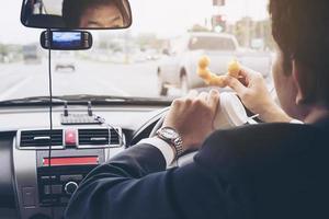 Mann, der Donuts mit Kaffee isst, während er Auto fährt - Multitasking-unsicheres Fahrkonzept foto