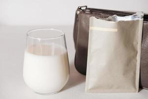 Kollagenpulver-Reisepackung und ein Glas Milch, um einen Proteincocktail zuzubereiten. Kollagenpeptidpulver für ein gesundes Getränk. Unterstützung für schöne Haut. Damentasche, um ein Proteinsäckchen für unterwegs mitzunehmen foto