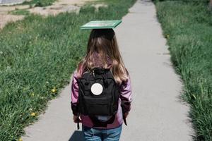 kleines Mädchen, das mit Schultasche und einem Buch auf dem Kopf zur Schule geht. foto