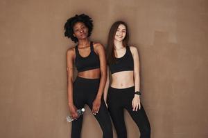Entspannung nach dem Fitnesstraining. Zwei multiethnische Freundinnen stehen im Studio mit braunem Hintergrund foto