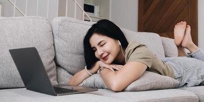 asiatische frau, die mit geschlossenen augen auf dem sofa im wohnzimmer liegt und musik auf einem laptop hört. Frau, die sich zu Hause auf dem Sofa entspannt, zeigt Nachrichten auf dem Computerbildschirm des Geräts an. foto