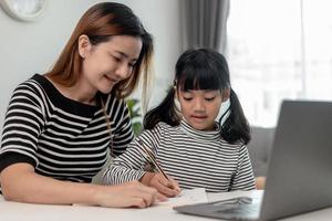 asiatisches kleines junges mädchen, das zu hause mit der mutter online-unterricht lernt. vorschulkind benutzt laptop-computer, macht hausaufgaben, homeschool vom schullehrer über digitales remote-internet mit unterstützung von mama. foto