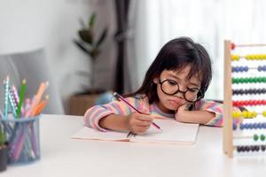 Kleines asiatisches Mädchen, das allein sitzt und mit gelangweiltem Gesicht herausschaut, Vorschulkind, das den Kopf auf den Tisch legt, mit trauriger Langeweile bei den Hausaufgaben, verwöhntes Kind foto