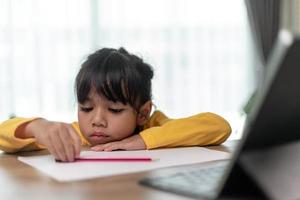Kleines asiatisches Mädchen, das allein sitzt und mit gelangweiltem Gesicht herausschaut, Vorschulkind, das den Kopf auf den Tisch legt, mit trauriger Langeweile bei den Hausaufgaben, verwöhntes Kind