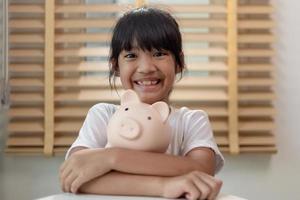 kleines asiatisches mädchen, das geld in einem sparschwein spart, über sparen lernt, kind spart geld für zukünftige bildung. geld, finanzen, versicherungen und personenkonzept foto