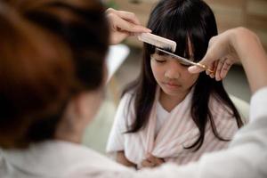 asiatische mutter schneidet ihrer tochter im wohnzimmer zu hause die haare, während sie während der sperrung zu hause sicher vor dem covid-19-coronavirus bleibt. selbstquarantäne und soziales distanzierungskonzept. foto