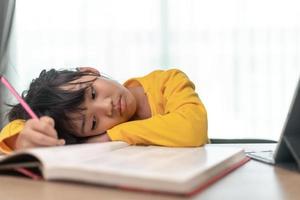 Kleines asiatisches Mädchen, das allein sitzt und mit gelangweiltem Gesicht herausschaut, Vorschulkind, das den Kopf auf den Tisch legt, mit trauriger Langeweile bei den Hausaufgaben, verwöhntes Kind foto