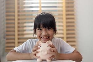 kleines asiatisches mädchen, das geld in einem sparschwein spart, über sparen lernt, kind spart geld für zukünftige bildung. geld, finanzen, versicherungen und personenkonzept foto