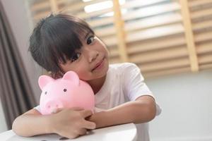 kleines asiatisches mädchen, das geld in einem sparschwein spart, über sparen lernt, kind spart geld für zukünftige bildung. geld, finanzen, versicherungen und personenkonzept foto