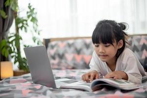 Kleines asiatisches Mädchen, das einen Laptop benutzt, während es auf dem Bett liegt foto