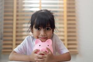 kleines asiatisches mädchen, das geld in einem sparschwein spart, über sparen lernt, kind spart geld für zukünftige bildung. geld, finanzen, versicherungen und personenkonzept foto