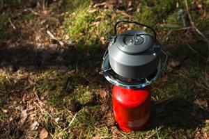 Kochen, Erhitzen eines touristischen Wasserkochers auf einem tragbaren Gasbrenner mit einer roten Gasflasche. Camping, ein Mann kocht das Frühstück im Freien. Sommeraktivitäten im Freien foto