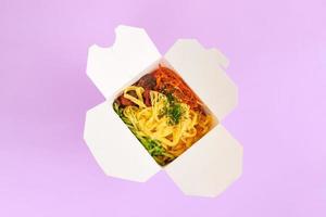 Reiswok mit Meeresfrüchten und Gemüse in weißer Box isoliert auf violettem Hintergrund, Fast-Food-Lieferung foto