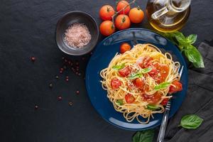 Leckere appetitliche klassische italienische Spaghetti-Nudeln mit Tomatensauce, Käse, Parmesan und Basilikum auf Teller und Zutaten zum Kochen von Nudeln auf dunklem Tisch. flache Ansicht von oben.