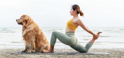 asiatische junge Frau beim Yoga mit ihrem Hund auf einer Yogamatte am Strand. Foto in Bannergröße