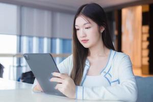 schöne junge asiatische berufstätige frau mit langen haaren im weißen hemd hält tablette, die sie glücklich lächelt, während sie auf stuhl im arbeitsbüro mit verglastem gebäude als hintergrund sitzt. foto