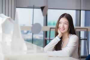 asiatische junge frau mit langen haaren lächelt, während sie in die kamera schaut, während sie auf einem stuhl am arbeitsplatz mit einem verglasten gebäude als hintergrund sitzt. foto