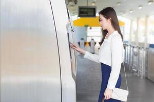 Schöne junge asiatische Geschäftsfrau in einem weißen Hemd steht, um einen Ticketautomaten in einer Skytrain-Station zu drücken, um im Büro zur Arbeit zu gehen. foto