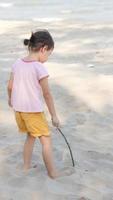 Positives charmantes 4-jähriges süßes asiatisches Mädchen, kleines Vorschulkind, das an einem schönen sonnigen Tag im Sommer am Sandstrand mit kleinem Holzstab schreibt und zeichnet. foto
