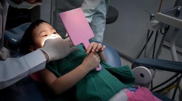 charmantes 3 Jahre altes kleines asiatisches Mädchen auf dem Zahnarztstuhl hält den Spiegel für die Zahnuntersuchung. foto