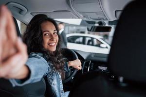 muss diesen Moment festhalten. süßes mädchen mit schwarzen haaren, das ihr brandneues teures auto im autosalon probiert foto