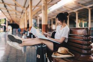 glückliche junge asiatin reisende oder rucksacktouristen, die karte verwenden, wählen, wohin sie mit gepäck am bahnhof reisen möchten, sommerferienreisekonzept foto