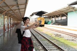 Schöne junge Asiatin mit einem Rucksack-Selfie mit Smartphone, während sie in der Nähe des Eisenbahnzuges auf dem Bahnsteig steht. Reisesommerkonzept foto