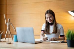 lächelnde Studentin trägt Notizen für das Online-Studium mit dem Lehrer, glückliche junge Frau lernt Sprache, hört Vorlesung, sieht sich das Webinar an, schreibt Notizen, schaut auf den Laptop zu Hause, Fernunterricht. foto