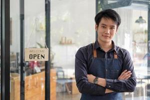 intelligenter asiatischer junger barista-mann in schürze, der tablette hält und vor der tür des cafés mit offenem schild steht. Unternehmer-Startup-KMU-Unternehmerkonzept foto