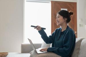 schöne asiatische frau, die mit laptop auf der couch im wohnzimmer sitzt und etwas aufregendes interessantes im fernsehen sieht foto