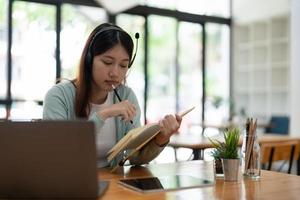 asiatische frau, die schreibt, macht liste, die sich notizen im notizblock macht, der drinnen auf dem laptop arbeitet oder lernt - bildungskurs oder training, seminar, bildungs-online-konzept foto