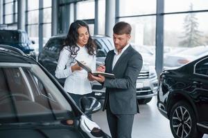 wo ich unterschreiben muss. Kundin und moderner stylischer bärtiger Geschäftsmann im Autosalon foto