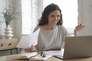 Fröhliche lächelnde weibliche Mitarbeiterin im Home Office mit Kopfhörern, die einen Videoanruf auf dem Laptop hat foto