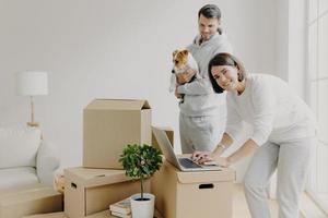 Glückliche Hauskäufer posieren neben ausgepackten Kartons, freuen sich über den Umzug in ein neues Haus, Frau sucht nach Ideen für die Renovierung des Schlafzimmers, Mann trägt Rassehund, beschließt, die Wohnung zu reparieren. Hypothekenkonzept foto