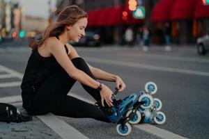 Außenaufnahme einer aktiven Frau, die Inline-Skates schnürt, bereitet sich auf die Fahrt vor, sitzt auf der Straße vor geschäftigem Stadthintergrund, gekleidet in schwarze Sportkleidung, genießt Rollschuhlaufen. sportliches lifestyle- und erholungskonzept foto