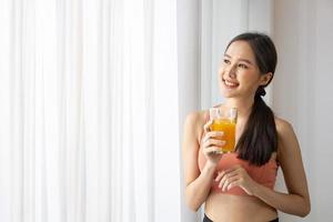 gesunde asiatin, die ein glas orangensaft zur erfrischung trinkt, während sie sportkleidung für ernährung und vitamin-c-ergänzungen am fenster mit kopierraum trägt foto