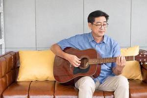 asiatischer älterer mann, der gitarre auf dem sofa innerhalb des hauses spielt. aktiver seniorenlebensstil nach der pensionierung mit glück und guter psychischer gesundheit. foto