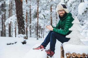 Foto von fröhlichen männlichen Typen Nachrichten auf dem Smartphone schaut direkt in die Kamera, sitzt im weißen Winterwald, lächelt glücklich, genießt die Kommunikation, schaut sich Videos an, bewundert Landschaften