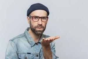 angenehm aussehender junger bärtiger mann mit schnurrbart, trägt modische brille, schwarzen hut und jeanshemd, bläst luftkuss in die kamera, drückt seine liebe zur freundin aus, isoliert auf grauem hintergrund foto