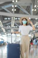 junge frau, die gesichtsmaske mit gepäck trägt, die auf dem flughafen spazieren geht, schutz-coronavirus-krankheitsinfektion, asiatische reisende mit hut. Reisezeit nach dem Impf-Booster-Dosierungskonzept foto