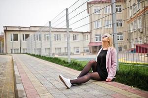 Blondes Mädchen mit Brille und rosa Mantel, schwarze Tunika, die auf der Straße am Zaun sitzt. foto