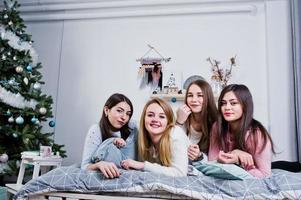 Vier süße Freundinnen tragen warme Pullover und schwarze Hosen auf dem Bett im neujahrsdekorierten Zimmer im Studio, spielen mit Kissen. foto