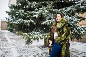 Brunettemädchen im grünen Pelzmantel am Wintertag gegen schneebedeckte Kiefer. foto