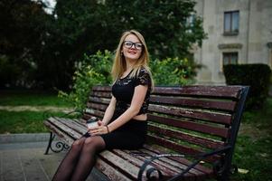 Elegantes blondes Mädchen auf Schwarz, das auf den Straßen der Stadt posiert und mit einer Tasse Kaffee in der Hand auf einer Bank sitzt. foto